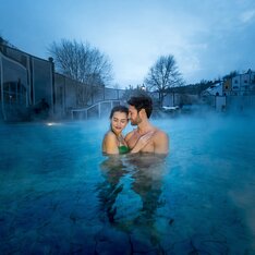 Paar entspannt im warmen Thermalwasser im Rogner Bad Blumau in der Dämmerung, umgeben von aufsteigendem Dampf – romantische Winterstimmung im Thermen- & Vulkanland Steiermark.